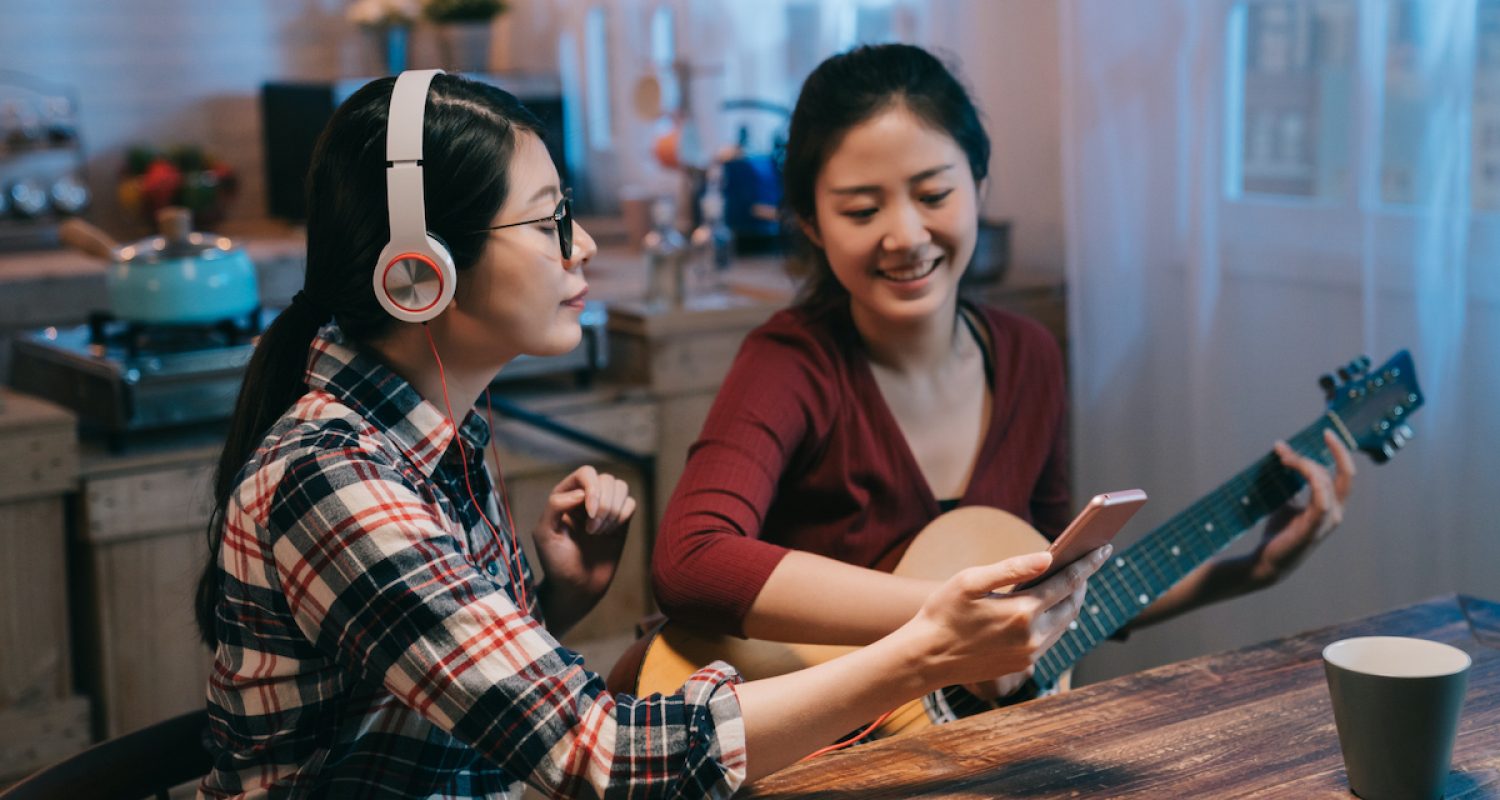 two asian japanese ladies friends sitting at wooden modern kitchen at home in midnight with guitar. young girl reading sheet music on cellphone. woman in headphones holding mobile phone enjoy melody